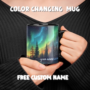 May include: A black color-changing mug with a design of a forest and aurora borealis. The mug has the text "your name" on the bottom. Free custom name.