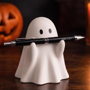 May include: A white ghost-shaped desk organiser with two small black eyes, holding a black ballpoint pen with silver accents and the text "F P E L Y W O O K" printed in white on the barrel. The ghost figure has a smooth, matte finish.