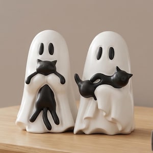 May include: Two white ghost figurines with smooth surfaces and black oval eyes. Each ghost is holding a small, glossy black cat figurine. The ghosts have rounded tops and draped, flowing edges at the base, creating a cohesive set of decorative items.