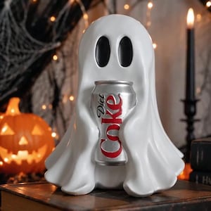 May include: A white ghost figurine with smooth, flowing fabric-like details and two large, oval black eyes. The ghost is holding a silver aluminium soda can with the red and white text Diet Coke printed vertically on the front of the beverage container.