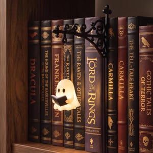 May include: A small, glowing, white ghost-shaped LED light holding a tiny black book, suspended by a silver chain from a black, ornate, wrought-iron style wall bracket mounted between a row of classic hardcover books on a wooden shelf.