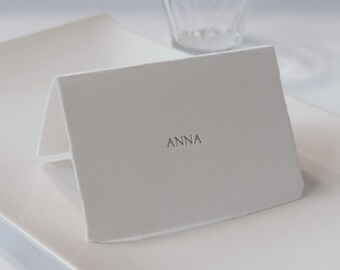 Puede incluir: Tarjeta de lugar blanca con el nombre "ANNA" impreso en letras doradas.