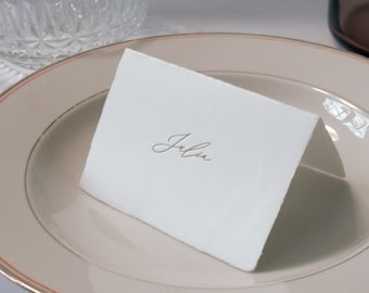 Puede incluir: Una tarjeta de lugar blanca con el nombre "Julia" escrito en cursiva. La tarjeta de lugar está sobre un plato beige con un borde dorado.