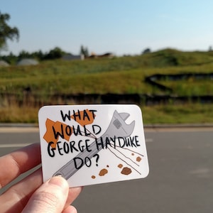 以下が含まれることがあります： 「What would George Hayduke do?」という黒い文字が書かれた白いステッカー。レンチと茶色の石もステッカーに描かれている。