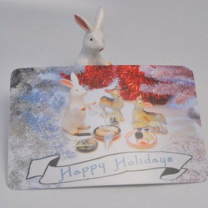 May include: A Christmas card with a white rabbit, two deer, and a white rabbit sitting at a table with food and drinks. The card has a silver and white background with red and white tinsel. The card says "Happy Holidays".