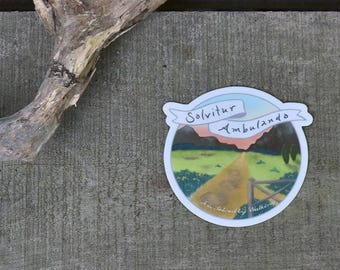 May include: A round sticker with a sunset scene and a road leading into the distance. The text "Solvitur Ambulando" is written on a banner above the scene. The text "As Solved By Walking" is written below the scene.