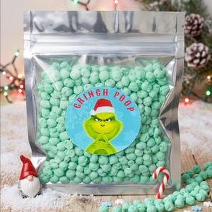 May include: A clear, resealable plastic pouch filled with small, light green, round sweets. A circular label on the front features a green character wearing a red Santa hat with the text "GRINCH POOP" printed in red letters above the character.