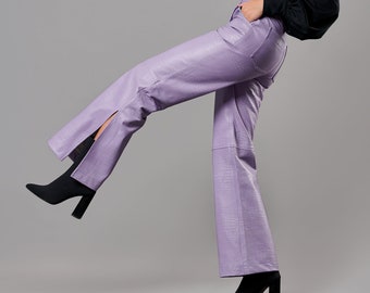 Può includere: Una donna indossa una camicia nera a collo alto e pantaloni di pelle viola con una fessura su una gamba. Indossa stivali neri con tacco e ha la mano in tasca.
