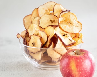 Op de afbeelding: Een glazen kom gevuld met gedroogde appelchips, met een hele rode appel naast de kom op een wit oppervlak.