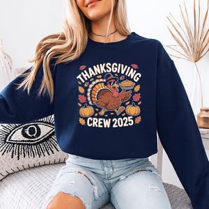 May include: Navy blue sweatshirt with a Thanksgiving-themed graphic. The graphic features a cartoon turkey wearing sunglasses and a chef's hat, holding a pie. The text "Thanksgiving Crew 2025" is displayed above and below the turkey.
