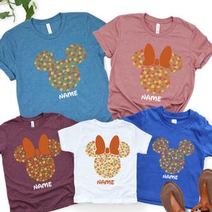 May include: Five short-sleeved t-shirts in various colors: teal, mauve, burgundy, white, and blue. Each shirt features a Mickey Mouse head design made of fall leaves and a bow, with the word "NAME" printed below.