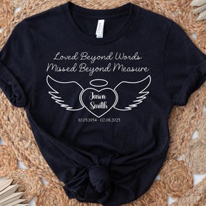 May include: A black t-shirt with white text and a heart-shaped design with wings. The text reads "Loved Beyond Words, Missed Beyond Measure." The heart contains the name "Jason Smith" and dates 10.05.1954 - 02.08.2025.