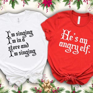 Peut inclure: Deux t-shirts, un blanc et un rouge, avec du texte sur le th&egrave;me des f&ecirc;tes. Le t-shirt blanc porte l'inscription "I'm singing I'm in a store and I'm singing" en script noir. Le t-shirt rouge dit "He's an angry elf" en script blanc. D&eacute;corations de vacances.