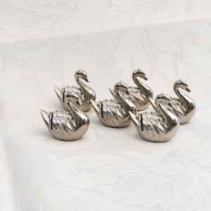 Peut inclure: Un ensemble de cinq petites figurines de cygnes en métal argenté. Chaque cygne est représenté dans une pose de nage avec des ailes texturées détaillées et un cou courbé. Les figurines sont disposées en groupe, révélant leur surface polie.