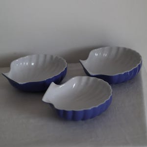May include: Three ceramic seashell-shaped bowls. The bowls have a white interior and a periwinkle blue exterior. The bowls are approximately 15 cm in diameter and are arranged on a white surface.
