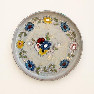 Può includere: Un piatto rotondo in ceramica con base grigio chiaro e motivo floreale dipinto a mano. Il design presenta un grappolo centrale di fiori rossi, blu e gialli circondato da un motivo circolare a tralci con fiori blu, gialli e foglie verdi.