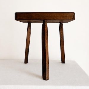 May include: A dark brown wooden stool featuring a flat, square top and three sturdy, tapered legs. The wood grain is visible across the surface, and the construction consists of a simple, functional design with a smooth, polished finish.