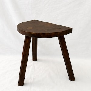 Puede incluir: Un taburete de madera marrón oscuro de tres patas con un asiento semicircular. Las patas están anguladas hacia afuera para mayor estabilidad. El taburete está hecho de madera maciza y tiene un aspecto rústico y artesanal. El asiento tiene un diámetro de unos 30 cm.