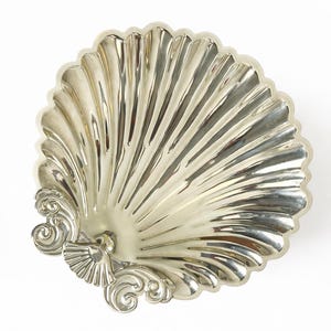 Puede incluir: Un plato decorativo plateado en forma de concha. El plato tiene un diseño detallado con estrías que irradia desde el centro, con adornos ornamentados y una pequeña figura de pájaro en la base. El plato está sobre un fondo blanco.