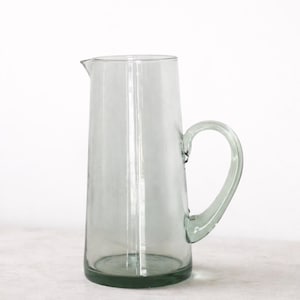 Könnte beinhalten: Eine transparente Glaskaraffe mit einem abgerundeten Griff und einem Ausguss. Die Karaffe hat einen leichten Grünton und steht auf einer hellen Oberfläche. Das Design ist schlicht und elegant, geeignet zum Servieren von Getränken. (Ca. 20 cm hoch)