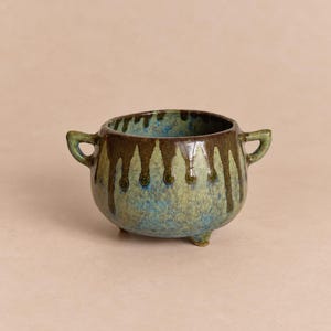 Pode incluir: Uma pequena tigela de cerâmica com duas alças laterais e três pés curtos. O exterior apresenta um esmalte azul e verde manchado com esmalte castanho escuro a escorrer da borda. O interior tem um acabamento em esmalte azul a condizer.