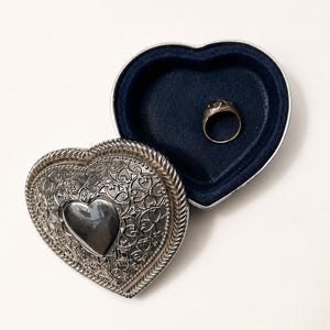 May include: A heart-shaped silver-toned metal trinket box with an intricate filigree pattern and a smooth central heart emblem. The lid is open, revealing a dark blue velvet-lined interior containing a single plain silver-toned metal ring.