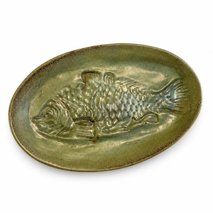 Peut inclure: Un plat de service ovale en céramique avec un motif de poisson en relief. Le plat est de couleur vert mat avec un bord plus foncé. Le poisson a des écailles et des nageoires détaillées, ajoutant de la texture. Un article unique et artisanal.