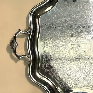 Peut inclure: Un plateau de service en métal argenté poli avec un bord festonné et une poignée décorative ornée d'un motif en coquille. La surface du plateau est décorée d'un motif complexe en relief représentant des volutes sur toute la zone centrale.