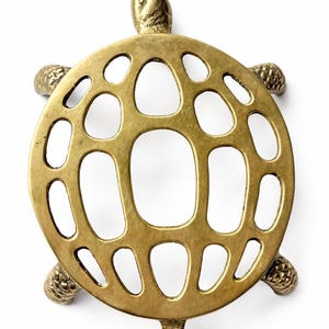 Puede incluir: Un salvamanteles de latón con forma de tortuga y un diseño calado decorativo. El salvamanteles tiene un caparazón redondeado con varias aberturas, cuatro patas y una cabeza y cola. El metal tiene un tono dorado cálido. Aproximadamente 15 cm de diámetro.