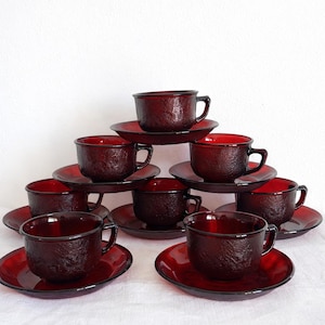 Può includere: Un set di dieci tazze da tè e piattini in vetro rosso rubino. Le tazze e i piattini sono disposti a forma di piramide. Le tazze hanno una superficie strutturata e un manico. I piattini sono rotondi e piatti.