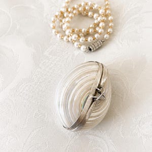 Peut inclure: Un récipient ovale en forme de coquillage irisé avec un fermoir en métal argenté et un collier de fausses perles avec des perles dorées. Les articles sont sur une surface blanche texturée.