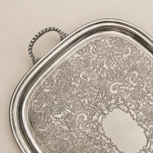 Peut inclure: Plateau de service argenté avec des détails floraux et des volutes ornés. Le plateau a une forme rectangulaire arrondie et une poignée décorative. La surface présente un cartouche vierge. Une pièce classique pour servir ou exposer.