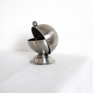 Puede incluir: Un azucarero pequeño, de color plateado, con tapa abisagrada y base redonda. La tapa está abierta, revelando el interior. El cuenco es de metal y está sobre una superficie blanca.