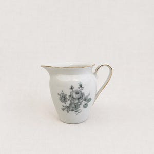 Peut inclure: Un pot à crème en porcelaine blanche avec un bord et une anse dorés. Le pot à crème présente un motif floral dans les tons de gris et de vert. Une pièce de vaisselle classique.