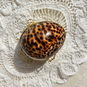 Puede incluir: Un pequeño bolso ovalado de concha con un marco y cadena de metal dorado. La concha tiene un patrón moteado marrón y beige. El bolso está sobre una superficie blanca y de encaje.