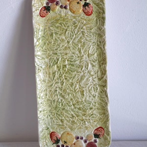 Può includere: Un vassoio rettangolare in ceramica verde chiaro con un motivo a foglie di felce strutturato. I bordi sono decorati con frutta in ceramica colorata, tra cui fragole, uva e pere. Il vassoio misura circa 36 cm di lunghezza.