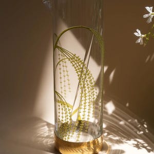 Peut inclure: Une carafe cylindrique haute en verre transparent ornée d'un motif botanique vert délicat représentant des tiges d'herbe tombantes. La base de la carafe est entourée d'une large bande dorée métallique avec un motif géométrique gravé.