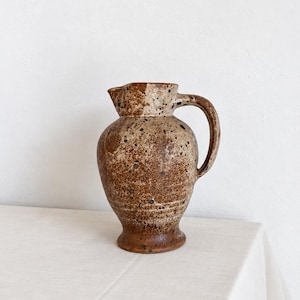 May include: A ceramic pitcher with a rounded body and a flared base, featuring a speckled brown and tan glaze. The vessel has a curved handle on the right side and a small pouring spout at the top rim, showcasing a textured, earthy appearance.