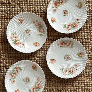 Peut inclure: Quatre assiettes blanches en forme de coquillage avec un motif texturé. Chaque assiette présente un motif floral avec des roses roses et oranges et des feuilles vertes. Les assiettes sont disposées sur une surface tannée tissée.