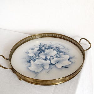 Peut inclure: Plateau de service rond avec bordure et poignées en laiton. Le plateau présente une surface blanche avec un motif de raisin et de feuilles bleu et blanc. Il repose sur de petits pieds en laiton.