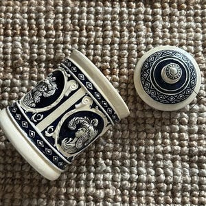Può includere: Un boccale da birra in ceramica color crema con coperchio, con elementi decorativi blu scuro e bianchi. Il boccale presenta due ritratti in rilievo dettagliati e motivi geometrici. Il coperchio è anch'esso color crema con un centro blu scuro e design geometrico.