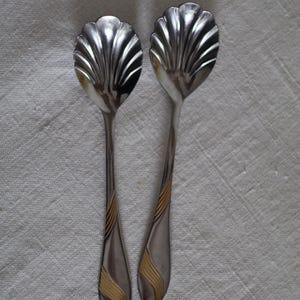 Pode incluir: Duas colheres de prata com uma tigela em forma de concha e um cabo longo. As alças têm um design ondulado dourado. As colheres são colocadas sobre uma superfície branca e texturizada.