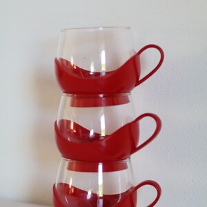 Puede incluir: Tres tazas de vidrio transparente apiladas, cada una con una base y un asa de color rojo vibrante. Las tazas tienen un diseño único, con la parte roja acunando el vidrio. Las asas son de color rojo a juego. Las tazas están apiladas.