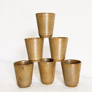 May include: A set of six stackable ceramic tumblers in a warm, earthy brown glaze. Each cup features a slightly tapered cylindrical shape with subtle horizontal ridges along the exterior surface, creating a consistent texture across the collection.