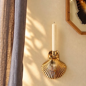 May include: A gold-colored metal wall sconce shaped like a scallop shell, featuring a central cylindrical holder for a single taper candle. The sconce is mounted on a plain wall, with a white taper candle inserted into the holder.