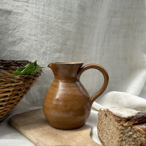 Puede incluir: Una jarra de cerámica marrón con asa, sentada sobre una tabla de cortar de madera con una rebanada de pan. La jarra está salpicada de manchas marrones y tiene un aspecto rústico.