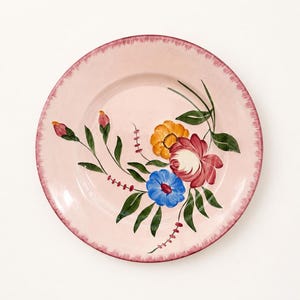 May include: A round ceramic plate with a light pink base color and a dark pink painted rim. The center features a hand-painted floral arrangement with a large red flower, a blue flower, a yellow flower, green leaves, and small red buds.