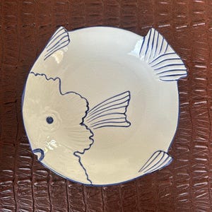 Puede incluir: Un plato de cerámica blanco con un diseño de pez. El plato presenta un contorno de pez azul claro con detalles azules, incluyendo aletas y cola. El plato es redondo y tiene un borde azul.