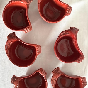 Peut inclure: Six plats de service en céramique rouge en forme de poulets. Chaque plat a une tête de poulet détaillée et des plumes de queue. Les plats sont d'un rouge profond et semblent être émaillés. Ils sont disposés sur une surface blanche.