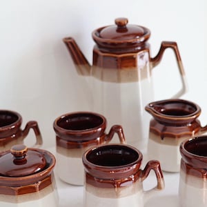Op de afbeelding: Een vintage keramische koffieset met een bruin en wit kleurenschema. De set bevat een theepot, een suikerpot met deksel, een melkkan en diverse mokken. De stukken hebben een geometrisch ontwerp en een glanzende afwerking, perfect voor koffie of thee.